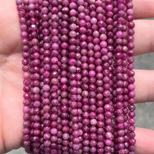 May include: A collection of multiple strands of small, round, faceted ruby beads. The beads are a deep, vibrant magenta-pink colour with a reflective, textured surface. Each bead is uniform in size and shape, arranged in parallel vertical rows.
