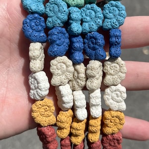 Peut inclure: Une collection de perles colorées en forme de coquillage, poreuses, dans des tons de vert, de bleu, de blanc, de jaune et de rouge. Les perles sont enfilées ensemble, créant un affichage vibrant et texturé. Les perles sont probablement en pierre de lave.