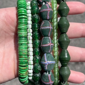 May include: A collection of green beaded bracelets in various styles. The bracelets feature round, disc, and geometric beads in shades of green, with some beads displaying red and white accents. The bracelets are held in a hand.