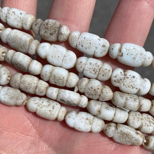 May include: A strand of white glass beads with a speckled, aged look. The beads are oval shaped and have a slightly irregular surface.