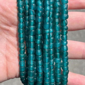 May include: A collection of multiple strands of small, round, translucent teal glass beads. Each bead features a slightly irregular, matte-textured surface, creating a uniform appearance across the vertical arrangement of the beaded strands.
