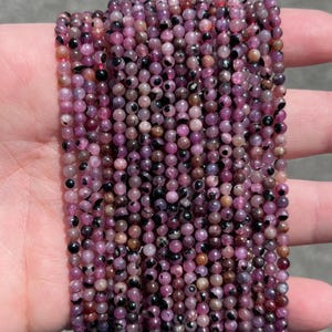 May include: A collection of small, round beads in pink, purple, and brown hues. The beads feature dark spots and are strung together. These beads are suitable for jewelry making. Each bead is approximately 0.3 cm in diameter.