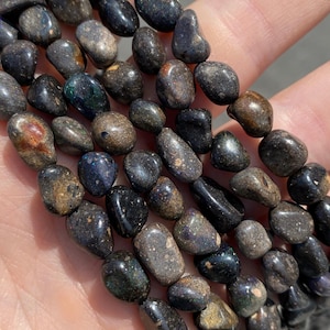 May include: Strand of polished, tumbled gemstone beads. The beads are a mix of dark colors, including black, brown, and gray, with iridescent flashes of blue and green. The beads are smooth and oval-shaped.