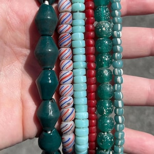 May include: A collection of beaded bracelets in various colors. The bracelets feature beads in shades of teal, red, white, and blue. Some beads are cylindrical, while others are faceted or round. The bracelets are held together by a string.