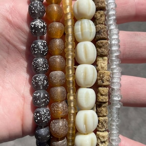 May include: A collection of beaded bracelets in various styles and colors. The bracelets feature beads in shades of black, brown, amber, cream, and clear. Some beads are round, others are textured or shaped like pumpkins. The bracelets are held in a hand.