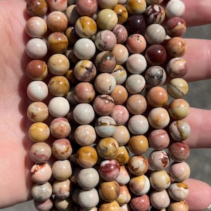 May include: A collection of round, polished gemstone beads arranged in multiple vertical strands. The beads feature a variegated colour palette of cream, tan, ochre, soft pink, and deep maroon, with natural veining and marbled patterns throughout.