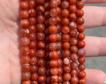May include: Strands of red and orange gemstone beads. The beads are round and have a polished, glossy finish. The beads are strung together, forming multiple strands. The color variations include shades of red, orange, and white.