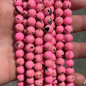 May include: A collection of round, bright pink beads arranged in multiple vertical strands. Each bead features a smooth, glossy finish with irregular black and metallic gold veining patterns distributed across the surface of the spherical shapes.