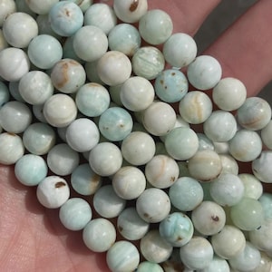 May include: A collection of round, light-colored beads with a mix of pale green, white, and brown hues. The beads are smooth and appear to be strung together, possibly for jewelry making or crafting. The overall aesthetic is natural and earthy.