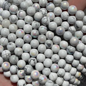May include: Close-up of a collection of white round beads with gray veining and iridescent sheen. The beads are smooth and appear to be made of a natural stone. Ideal for jewelry making or crafting.