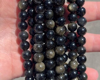 May include: A collection of polished, spherical beads arranged in multiple vertical strands. The beads are primarily black with shimmering, metallic gold inclusions visible within the stone, creating a reflective and textured appearance throughout.