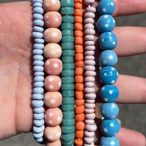 May include: A collection of colorful beaded bracelets. The bracelets feature a variety of colors, including light blue, peach with white polka dots, teal, orange, light pink, and blue with white polka dots. The beads are strung together to create a stylish accessory.