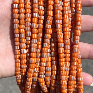 May include: A collection of multiple strands of small, cylindrical glass beads. Each bead features a vibrant orange base colour with distinct, vertical white stripes running along the length of the bead, creating a consistent pattern across all strands.