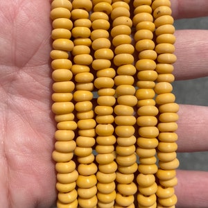 May include: Close-up of several strands of light yellow wooden beads. The beads are round with a slightly flattened shape, strung together to create a bracelet or necklace. The beads have a matte finish and a consistent color throughout.