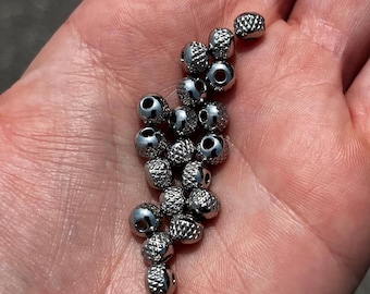 May include: A collection of small, silver-colored beads with a textured, diamond-like pattern. Each bead has a hole running through the center. The beads are shown in a person's open hand.