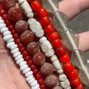 Op de afbeelding: Een verzameling kralenstrengen in verschillende kleuren en vormen. De kralen omvatten wit, rood en helder glas, evenals enkele met een getextureerd, verouderd uiterlijk. De kralen zijn er in verschillende maten en vormen, waaronder rond, cilindrisch en facetgeslepen.