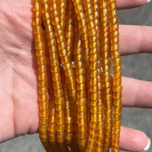 May include: Close-up of multiple strands of amber-coloured glass beads. The small, rectangular beads are strung together, creating a textured, flowing appearance. The beads have a warm, translucent quality, possibly vintage or handmade.