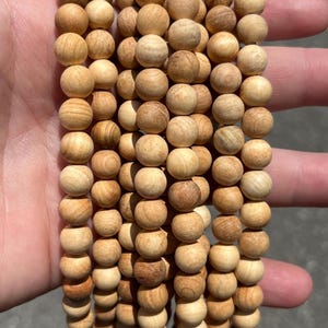 Puede incluir: Una colección de múltiples hilos de cuentas de madera redondas de color marrón claro. Cada cuenta presenta un patrón de grano natural con tonos variados de beige y marrón claro. Las cuentas son uniformes en tamaño y forma.