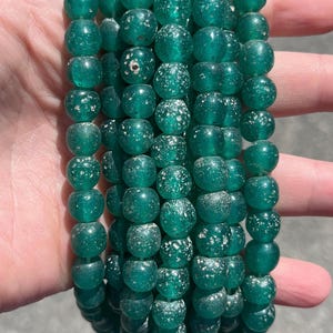 May include: Multiple strands of teal-coloured, translucent, round beads. The beads have an irregular shape and a speckled, aged appearance. The beads are strung together. The beads are held in a hand.