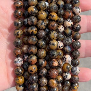 May include: A collection of spherical beads arranged in multiple vertical strands. The beads feature a mottled pattern with a mix of earthy brown, tan, black, and cream-colored spots, creating a natural, speckled appearance across the smooth surface.