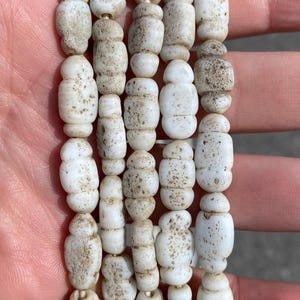 Puede incluir: Un collar de cuentas cilíndricas de color blanco roto con una superficie texturizada y porosa. Cada cuenta presenta una serie de crestas horizontales y está ensartada en un cordón fino, creando una forma alargada y uniforme con un aspecto mate.
