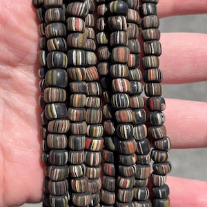 May include: Several strands of dark, barrel-shaped beads with a striped pattern in black, white, red, yellow, and brown. The beads are strung together, creating a textured and colourful display. The beads are held in a hand.