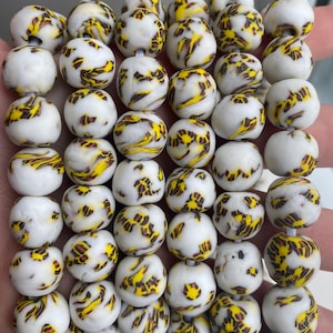 May include: A strand of white glass beads with yellow and brown swirling patterns. The beads are round and have a smooth, polished finish.