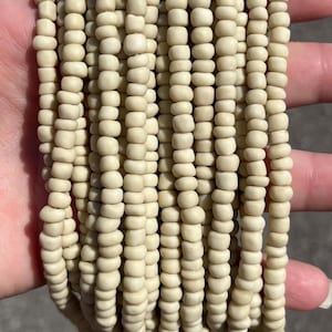 May include: A collection of multiple strands of small, round, off-white beads. Each strand consists of numerous uniform, matte-finish beads strung together in a vertical arrangement, creating a dense, textured appearance of light cream-colored units.
