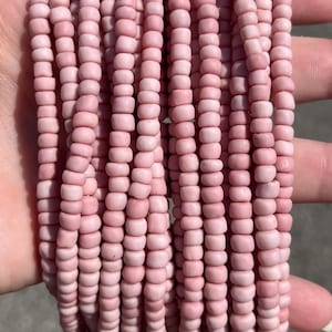 Könnte beinhalten: Eine Ansammlung mehrerer Stränge aus kleinen, runden Perlen mit mattem Finish in einem sanften, blassen Rosa. Die Perlen sind in Form und Größe einheitlich und in parallelen vertikalen Linien aufgereiht, was ein dichtes, strukturiertes und monochromes Bild ergibt.