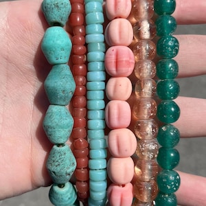 May include: A collection of beaded bracelets in various colors. The bracelets feature turquoise, red, light blue, pink, clear, and green beads. The beads have different shapes and textures, including round, faceted, and textured.