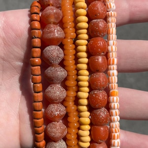 May include: A collection of beaded bracelets in shades of orange, yellow, and white. The bracelets feature various bead shapes, including round, cylindrical, and textured. The color palette ranges from light yellow to deep orange.
