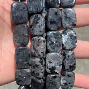 May include: A strand of rectangular, polished beads made of dark grey stone with black inclusions and subtle iridescent blue flashes. The beads are smooth, flat, and arranged in a vertical column, showcasing a natural mottled texture throughout.