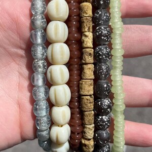 May include: A collection of beaded strands in various colors and textures. The strands include clear, white, brown, tan, black, and light green beads. The beads have different shapes, including round, cylindrical, and textured. The beads are held in a hand.