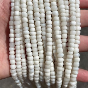 May include: A collection of multiple strands of small, opaque white beads. Each strand consists of numerous rounded, uniform beads strung together in a vertical arrangement, creating a dense, textured appearance with a consistent matte finish.