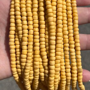 May include: A collection of multiple strands of small, round, matte yellow beads. The beads are uniform in size and shape, arranged in parallel vertical lines, creating a dense, textured appearance with a consistent mustard yellow colour throughout.