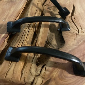 Op de afbeelding: Drie zwarte metalen kastgrepen met een vierkante vorm aan de uiteinden. De grepen zijn gemonteerd op een houten oppervlak.