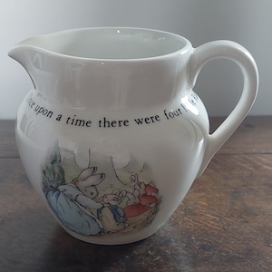 Può includere: Brocca in ceramica bianca con manico, con un'illustrazione di Beatrix Potter di quattro conigli in un ambiente boschivo. Il testo "Once upon a time there were four little rabbits" è stampato sul lato.