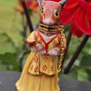 Op de afbeelding: Een keramieken beeldje van een muis in een gele jurk met rode bloemmotieven. De muis heeft een gouden kraag en ketting. De achtergrond toont rode bloemen en groen gebladerte.