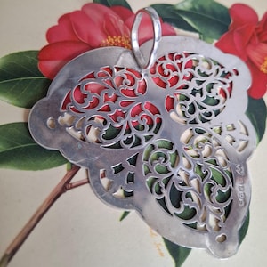 Op de afbeelding: Een zilverkleurige, sierlijke serveerschaal met een opengewerkt, bladvormig ontwerp. De schaal heeft een handvat en staat tegen een achtergrond van rode bloemen en groene bladeren. Het ingewikkelde patroon voegt een decoratief tintje toe.