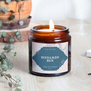 May include: A brown glass candle jar with a white label that says "Your Brand Cinnamon Bun Plant Wax Blend 11.5 oz". The candle is lit and the flame is visible.