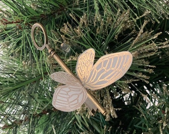 May include: A gold-colored key with a white and gold butterfly design ornament hanging on a green artificial Christmas tree.
