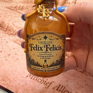 May include: A small, amber-colored glass bottle labeled "Felix Felicis" with gold accents and a cork stopper. The label reads "Liquid Luck" and "Mischief Alley Apothecary." A gold clover charm adorns the top. The bottle is held against a background with the words "Mischief Alley."