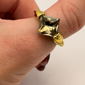 May include: A gold ring with a square, light brown gemstone. The ring's band has a textured design, and the gemstone is held in place by ornate gold accents. The ring is shown on a finger.