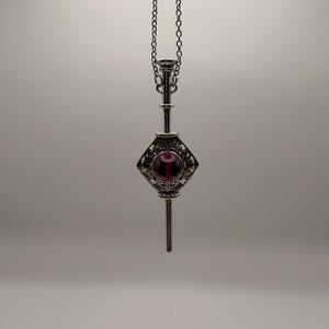 Könnte beinhalten: Eine silberfarbene Anhängerkette mit einem Schlüsselmotiv. Der Anhänger hat ein rautenförmiges Mittelstück mit einem verzierten Filigranmuster und einem roten Edelstein. Der Schlüssel hängt an einer zarten Kette.