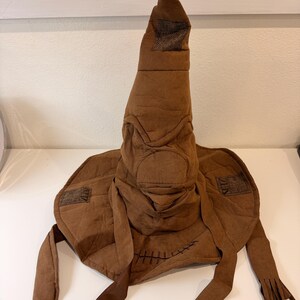 May include: A brown, fabric Sorting Hat with a tall, pointed top and a wide brim. The hat has a textured appearance, with patches and stitched details. Long, fringed ribbons hang from the sides. A classic costume accessory.