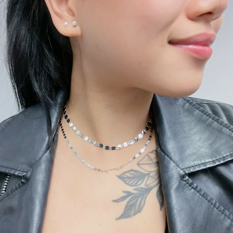 May include: Two silver chain necklaces with small, round, flat links. The top necklace has black links, and the bottom necklace has silver links. The necklaces are layered on a person's neck.