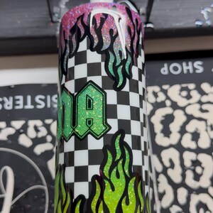 May include: A custom tumbler with a black and white checkered pattern, green and yellow flames, and the word "DA" in a green glitter font. The top of the tumbler has pink and black glitter flames.
