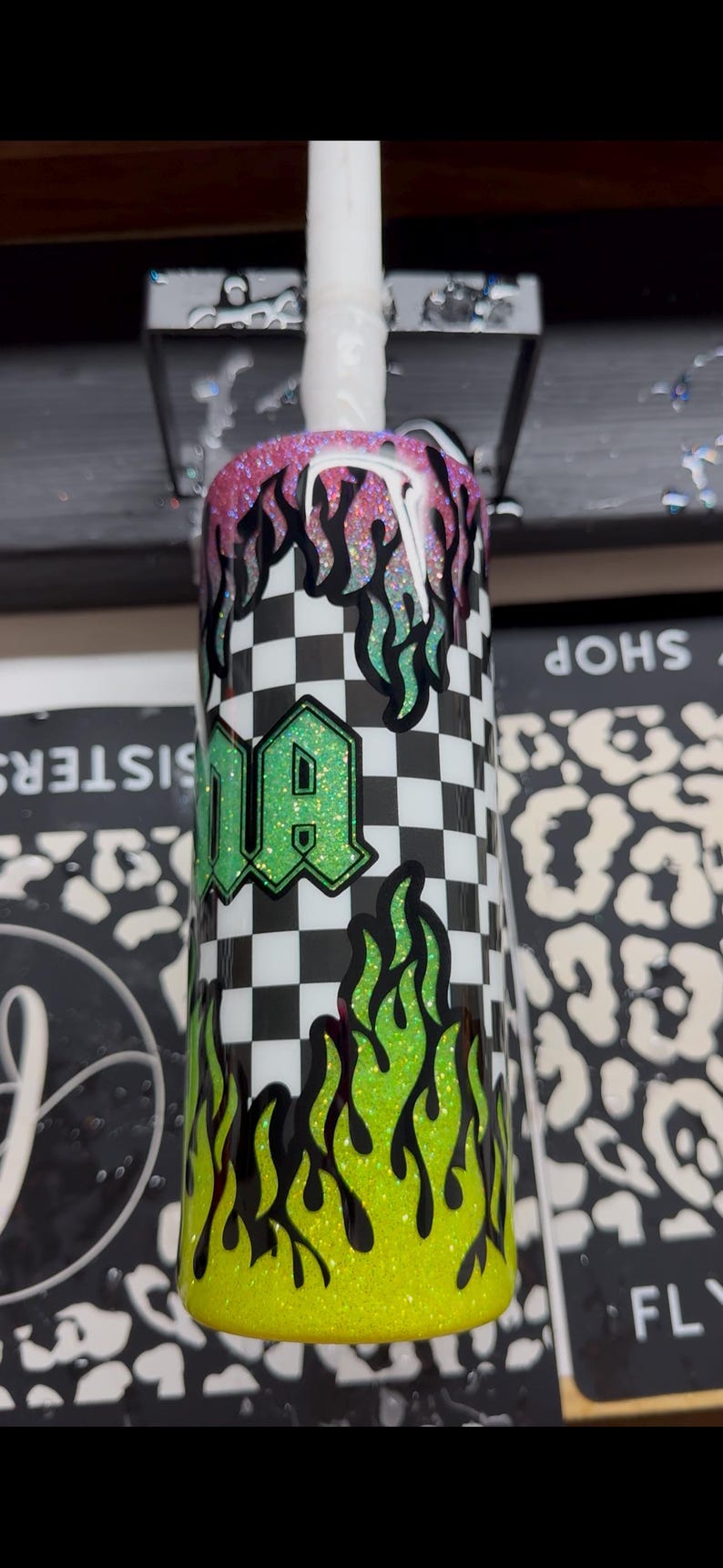 May include: A custom tumbler with a black and white checkered pattern, green and yellow flames, and the word "DA" in a green glitter font. The top of the tumbler has pink and black glitter flames.
