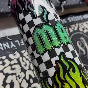 May include: A glittery tumbler with a black and white checkered pattern and green flames. The word "MQA" is displayed in a green glitter font. The top rim has pink glitter and the bottom has yellow glitter.