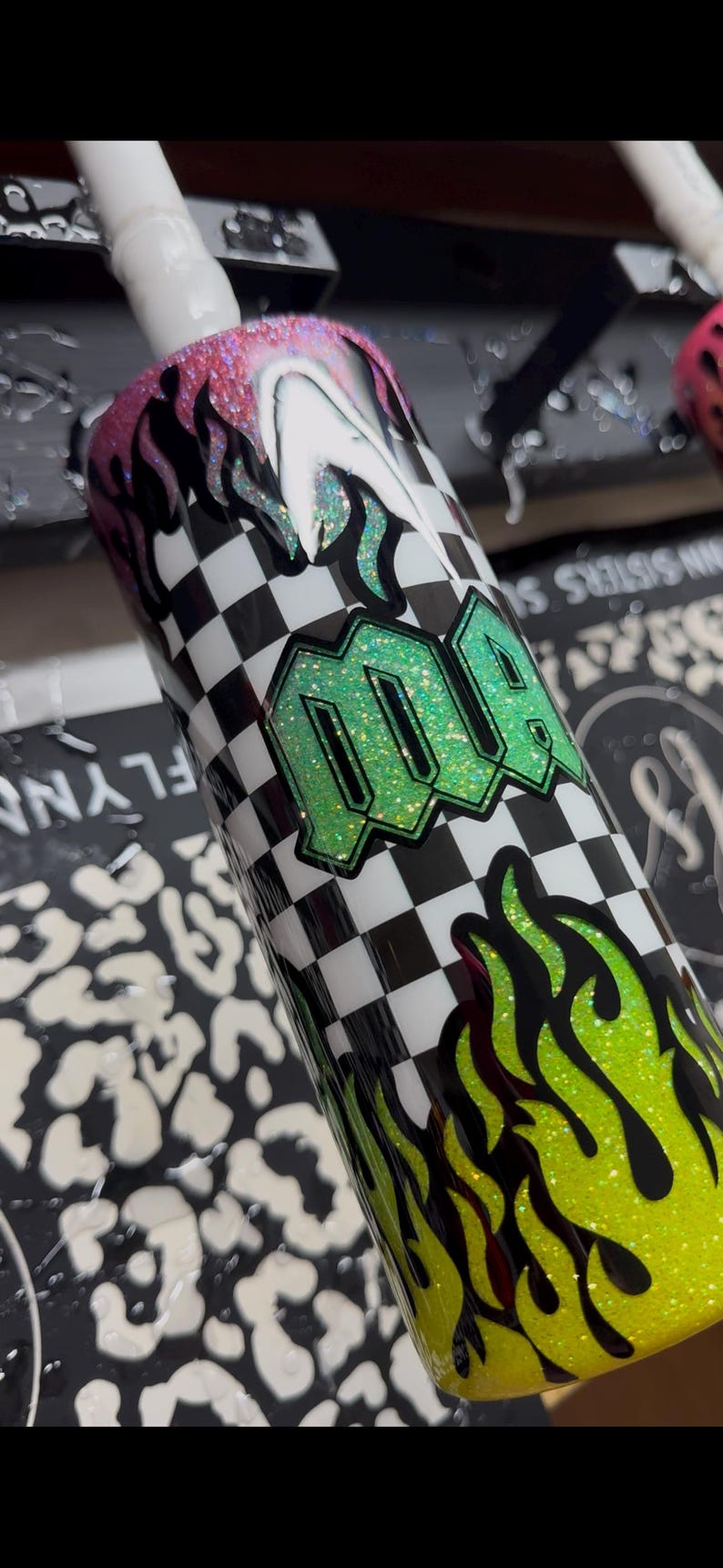 May include: A glittery tumbler with a black and white checkered pattern and green flames. The word "MQA" is displayed in a green glitter font. The top rim has pink glitter and the bottom has yellow glitter.